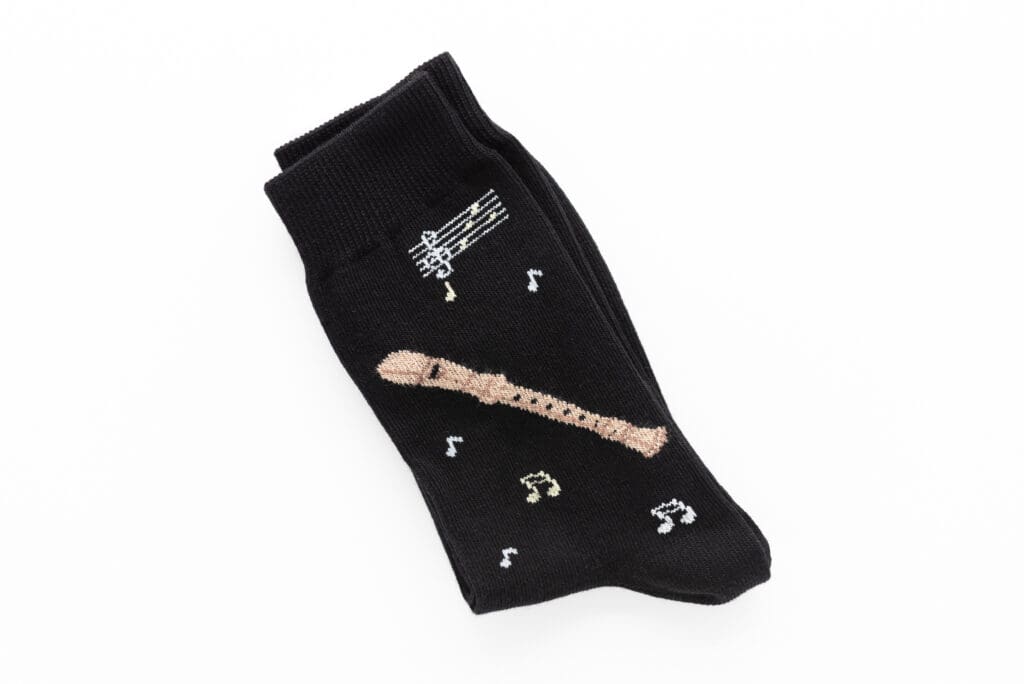 Schwarze Socke mit einem gestrickten hellbraunen Blockflöten Motiv, mehreren kleinen Musiknoten und einer weißen Notenzeile auf der Vorderseite.