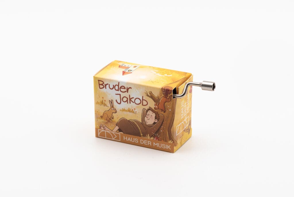 Handkurbel-Spieluhr mit gelb-brauner Illustration eines liegenden Mönchs unter einem Baum, einem Hasen und dem Text "Bruder Jakob" sowie Haus der Musik.