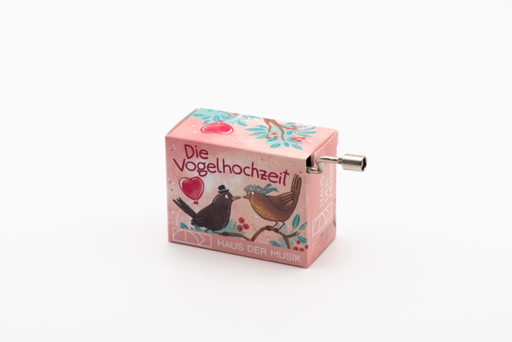 Rosafarbene Handkurbel-Spieluhr mit der Aufschrift "Die Vogelhochzeit" und einer Illustration zweier Vögel auf einem Zweig mit Herzmotiven.