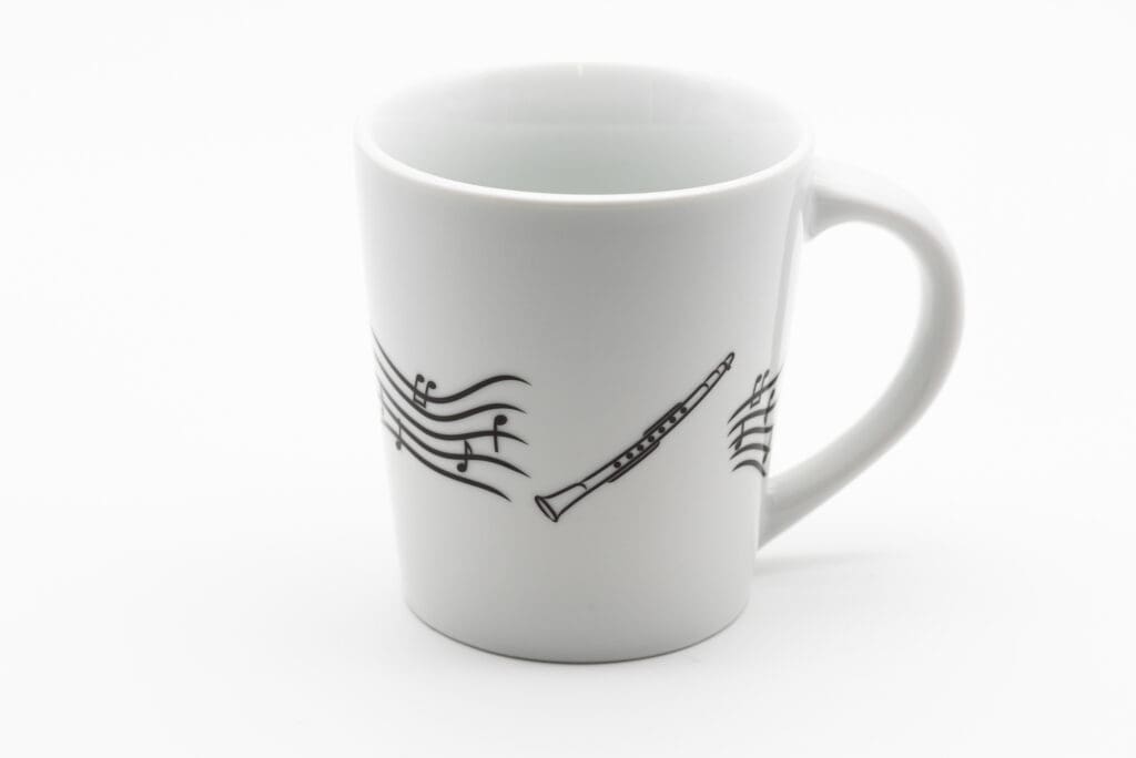Weiße Tasse mit schwarzer Illustration einer Klariette und umliegenden Notenlinien auf glatter Oberfläche.
