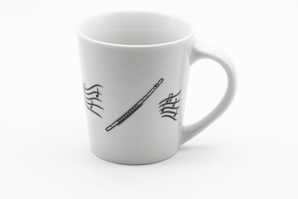 Weiße Tasse mit schwarzer Illustration einer Querflöte und umliegenden Notenlinien auf glatter Oberfläche.