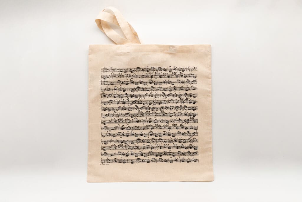 beigefarbene Tasche mit langen Henkeln und schwarzem Aufdruck einer mehrzeiligen handgeschriebenen Musikpartitur.